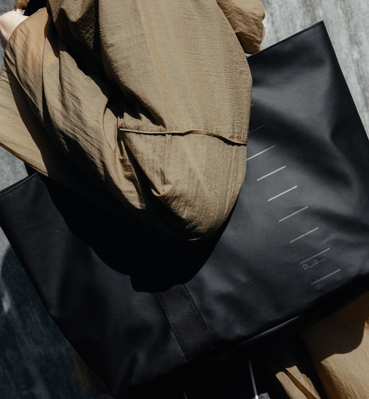 Dbブランドの軽量トートバッグ「Essential Tote 16L」のモデル着用画像。旅行やスポーツを楽しむアクティブな男女に最適な、シンプルで収納力に優れた自立式デザインです。色はBlack Out
